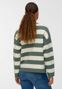 Donna con coda di cavallo riccia, che indossa una maglietta a maniche lunghe verde e bianca a righe e jeans blu, di spalle contro uno sfondo neutro.