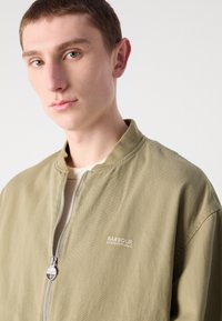 Olijfgroene bomberjack met een ritssluiting aan de voorkant, geribbelde kraag en het "Barbour International" logo in het wit op de linkerborst. Glad textuur.