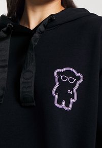 Schwarzer Kapuzenpullover mit einem Bärenmotiv, der eine Brille trägt, und "GA" auf der Brust in weißer Umrandung, mit strukturierten schwarzen Kordeln.