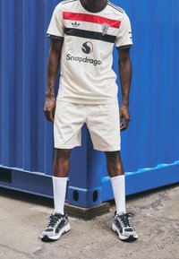 Valkoinen urheilupaita mustilla ja punaisilla raidoilla, beigehousut ja harmaat lenkkarit. Mallilla on valkoiset sukat ja hän seisoo sinistä taustaa vasten.