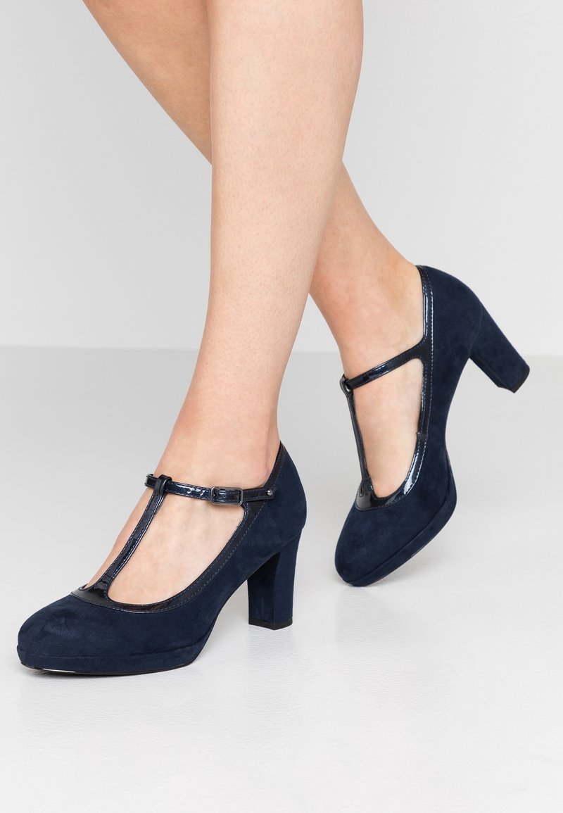 Pieds chaussés de chaussures en daim bleu marine à bride en T et talons moyens, sur un fond uni gris clair.