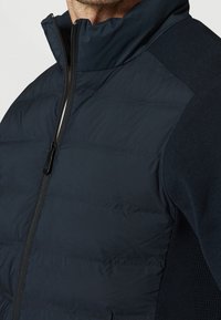 Chaleco acolchado azul marino con cuello alto, con cremallera en la parte delantera y lados de punto texturizados para mayor elasticidad y transpirabilidad.