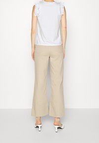 Biała bezrękawnik z falbanami na ramionach, połączona z beżowymi spodniami zestawionymi w szerokie nogawki i białymi klapkami na obcasie. Gładki materiał i prosty design.