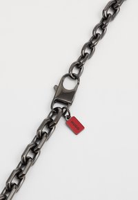 Kraftig metal kæde i mørkegrå med en blank finish. Har en sølvfarvet lås og et lille rødt mærke med teksten "HUGO."