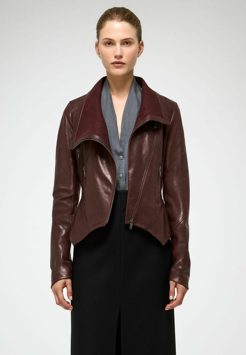 Veste en cuir bordeaux avec un large col, fermeture éclair à l'avant et un design épuré. Associée à une chemise grise et une jupe noire, avec des accents matériels minimalistes.