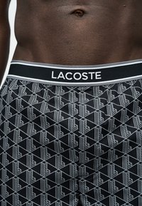 Μαύρα και λευκά σορτς με γεωμετρικό σχέδιο και μαύρη μέση με τη λέξη «LACOSTE», φορεμένα από άτομο με σκουρόχρωμο δέρμα.