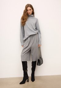 Pelēks adīts turtlenecks džemperis, pelēki pielāgoti šorti, melnas augstās zābaki līdz ceļiem ar rāvējslēdzēju un melna quilted rokas soma ar ķēdes siksnu.