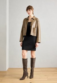 Veste en cuir marron cropped avec poches, robe noire ajustée et bottes à talons aiguilles léopard jusqu'aux genoux sur un sol en bois.