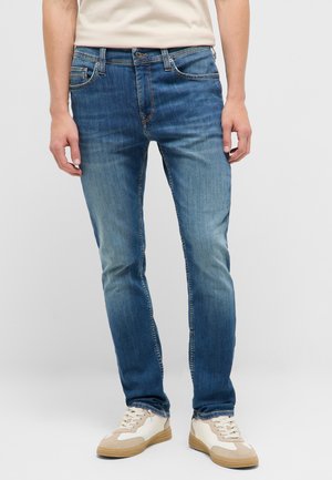 Man wearing blue slim-fit jeans with fading details and white sneakers, standing against a plain light background.