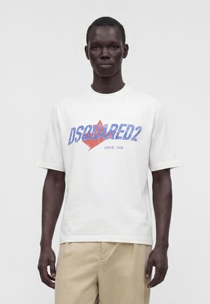 Mann mit dunkler Haut, der ein weißes DSQUARED2 T-Shirt mit blauem und rotem Logo und beigen Hosen trägt, stehend vor einem einfarbigen weißen Hintergrund.
