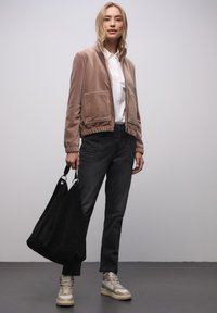 Světle hnědá velurová bomber bunda se dvěma předními kapsami, kombinovaná s černými džínovými kalhotami a béžovými kotníkovými teniskami. V ruce držena černá kabelka přes rameno.