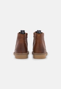 Botas de piel marrón hasta el tobillo con costuras verticales, superficie texturizada y suela de goma plana. Presentan una lengüeta y acentos amarillos.