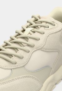 Béžové tenisky s kombináciou hladkých a semišových textúr, zaobleným tvarom, polstrovaným jazykom a zodpovedajúcimi šnúrkami. Jemný modrý akcent.