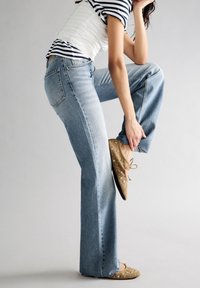 Mujer vistiendo unos vaqueros de pernera ancha en dos tonos y unas planas marrones con tachuelas, levantando una pierna y sosteniendo su tobillo contra un fondo claro y liso.