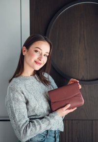 Burgunderfarbene Lederhandtasche mit Klappendesign und geprägtem Logo, gehalten von einer Person in einem hellgrauen Strickpullover vor einem dunklen Holz-Hintergrund.