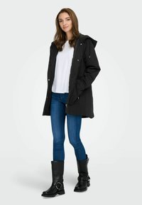 Črna kapuca parka z zadrgo, v kombinaciji z belo srajco in modrimi kavbojkami. Model nosi črne visoke škornje do kolen z zaponkami.