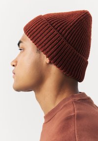 Rostu boje pleteni šešir s rebrastom teksturom, koji se sužava prema vrhu ušiljenom u točku. Nošen ukoso, ističući glatku kožu ispod.