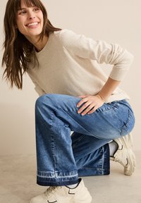 Beige Strickpullover, kombiniert mit hellblauen weit geschnittenen Jeans, die am Saum einen Batikakzent aufweisen, abgerundet mit klobigen weißen Sneakers.