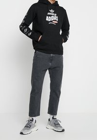 Černá mikina Adidas s bílým logem a textem, v kombinaci s tmavě šedými džíny a šedobílými teniskami, stojící na osobě proti jednobarevnému pozadí.