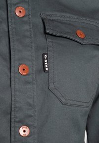 Dark gray shirt made of textured fabric, featuring copper-colored buttons, a chest pocket, and a black G-STAR label.