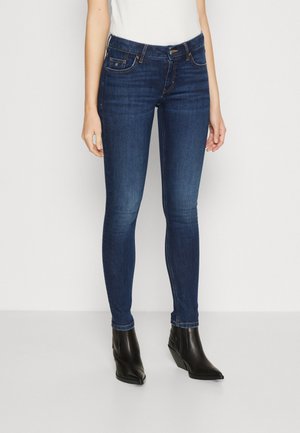 Skinny blue jeans in denim fabric with a mid-rise waist, five-pocket design, and visible stitching details, worn with black ankle boots.
