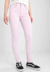 Jeans skinny rosa claro con una textura suave y diseño de cinco bolsillos, combinados con zapatillas negras y blancas que presentan un acento a rayas.