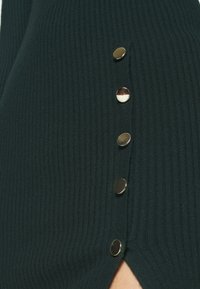 Rippender, dunkelgrüner Stoff mit seitlichem Schlitz, der mit fünf goldfarbenen Knöpfen verziert ist. Die Textur ist weich und betont die körpernahe Passform des Kleidungsstücks.