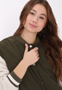 Olive zöld ribbed dzseki fehér bolyhos béléssel, patentos záródással és csíkos mandzsettával. A modell az egyik kezével a gallért tartja.