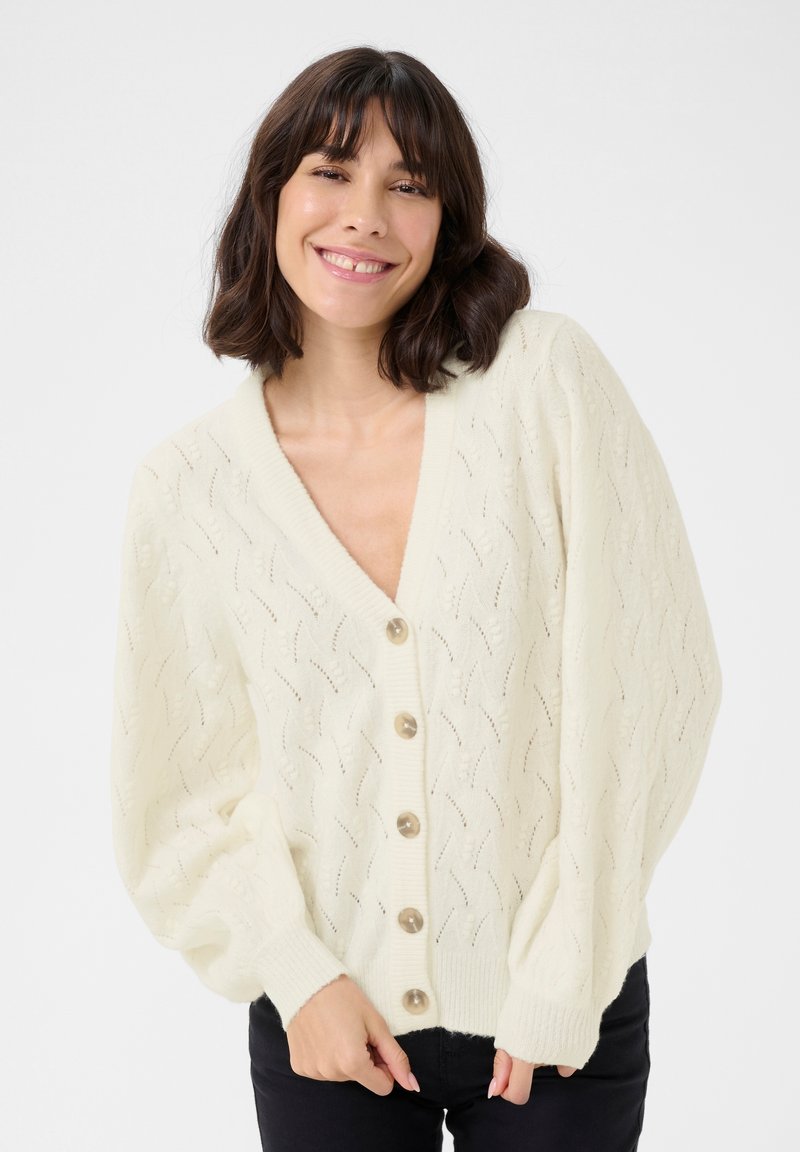 Cream knit cardigan with a V-neck, button front, and textured pattern. Features puffed sleeves and five round, light brown buttons.