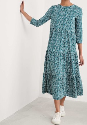 Mujer con vestido midi color teal con patrón blanco de hojas y mangas tres cuartos, de pie con una mano apoyada en una pared blanca, zapatillas blancas.