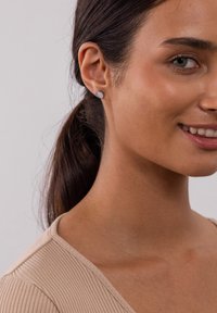 Graue Ohrstecker in kreisförmigem Design, am linken Ohr getragen. Glatte Textur, minimalistischer Look. Helle Hautfarbe mit zurückgebundenem dunklem Haar.