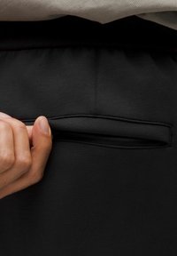 Zwarte sportbroek heeft een stijlvolle uitstraling met een verborgen zak, een soepele textuur en een elastische tailleband. Een hand wordt getoond die de zak opent.