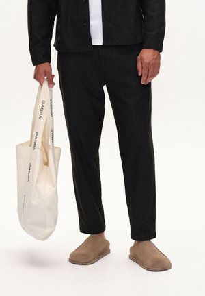 Mann trägt schwarze Hose, schwarze Jacke, beige Slipper, hält weiße Tragetasche mit schwarzem "GABBA"-Schriftzug auf den Trägern und Seiten.