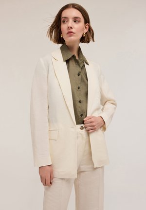 Blazer en lin crème avec des revers structurés, associé à une chemise boutonnée en olive texturé, stylé avec un pantalon neutre.
