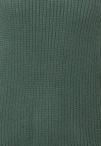 Pletený svetr v tlumené zelené barvě. Má žebrovanou strukturu a přiléhavý střih, který zdůrazňuje teplo a univerzálnost.