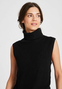 Melna bez piedurknēm turtlenecka blūze ribiņas struktūrā, ar augstu apkakli un vienkāršu dizainu, uzsverot komfortu un daudzpusību.