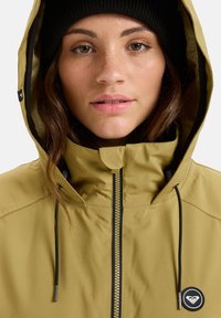 Svetlo rjava vodoodporna jakna z zadrgo, nastavljivo kapuco in črnimi vrvicami. Ima okroglo logo na prsih.