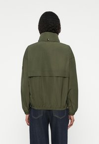 Ελιά-πράσινο μπουφάν με ψηλό γιακά, ελαστική μέση και άνετη εφαρμογή. Διαθέτει ένα καπάκι στο πίσω μέρος και κουμπιά διακοσμητικά στον γιακά.