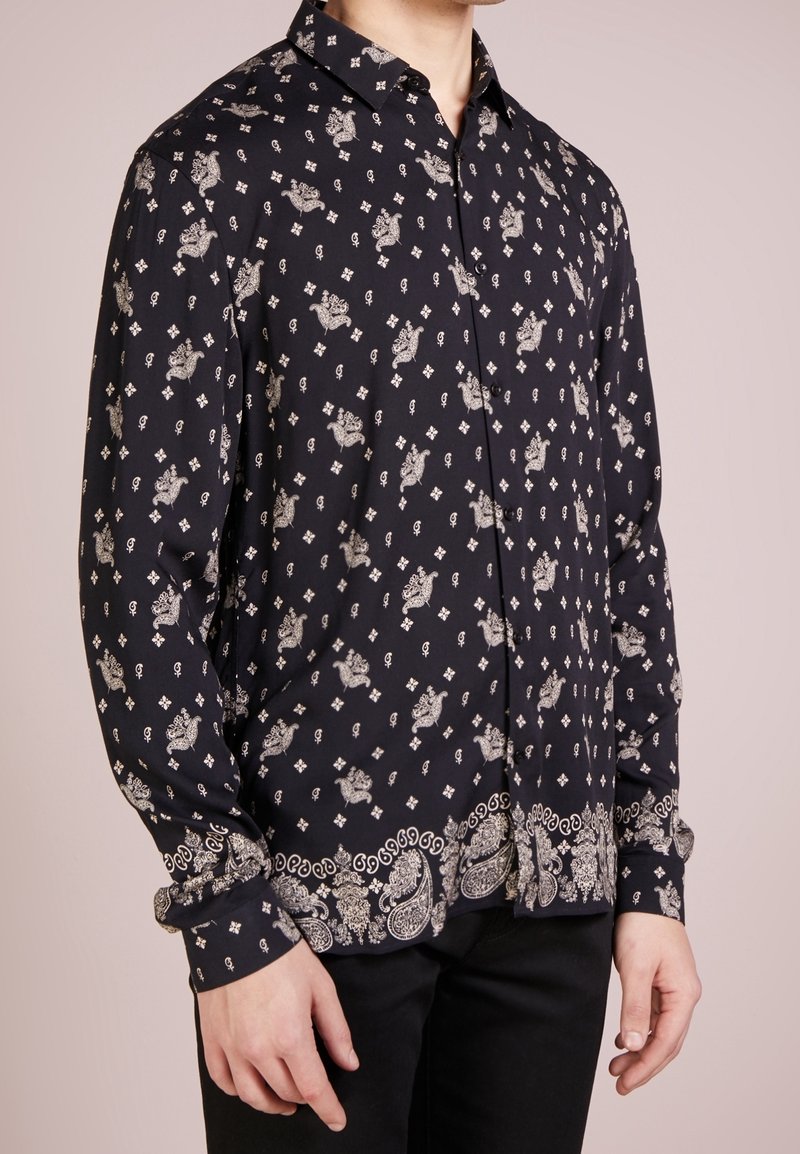 Homme portant une chemise noire à manches longues boutonnée avec des motifs floraux et cachemire blancs, associée à un pantalon noir, debout devant un fond uni.