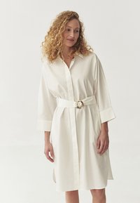 Valge nupuja dress, mis on valmistatud siledast kangast, rõhutatud vöökohaga, millel on ümmargune kuldne pandla ja kolmveerandi varrukad.