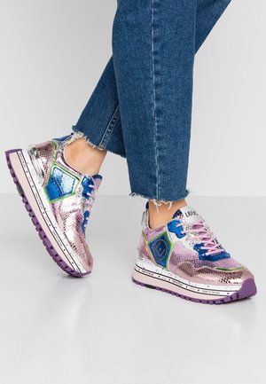 Stopy w błyszczących różowych, niebieskich i zielonych sneakersach ze skóry węża z fioletowymi podeszwami i sznurowadłami, zestawione z dżinsami o postrzępionych nogawkach.