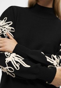 Pull noir à manches longues avec broderie florale blanche sur les manches, poignets côtelés et col montant, porté par une personne croisant les bras.