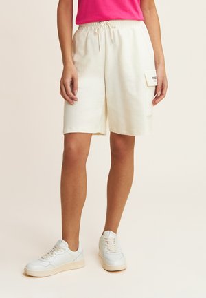 Pantaloni corti bianchi in cotone con vita elasticizzata, cordino regolabile, tasche laterali e una piccola toppa sul lato. Abbinati a sneakers chiare.