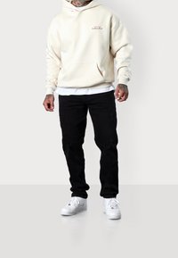 Krémová mikina s kapucí a přední kapsou, v kombinaci s roztrhanými černými džínami. Outfit doplňují bílé tenisky a vrstvená košile pod mikinou.