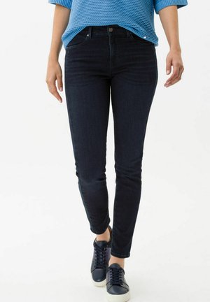 Jean skinny bleu foncé avec une texture lisse, doté d'une fermeture à bouton à l'avant et de poches latérales, associé à des baskets bleu marine.