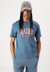 Blauwe katoenen T-shirt met "MALIBU" in het rood gedrukt, gecombineerd met bijpassende blauwe joggingbroek. Grijze pet met diverse decoratieve speldjes.
