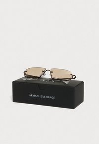 Pruunikate ristkülikukujulised prillid õhukeste metallraamidega, eksponeeritud musta kasti peal, millel on kiri "Armani Exchange". Selged tempelaktsendid on nähtavad.