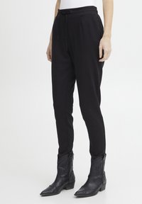 Persona che indossa pantaloni neri affusolati con tasche laterali e stivaletti alla caviglia in pelle nera stile western su sfondo neutro.