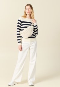 Maglione a righe crema e blu navy con polsini a costine, abbinato a pantaloni bianchi larghi e a una borsa a tracolla beige e pelosa. Sneakers bianche completano il look.