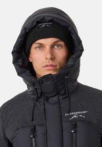 Black puffer jacket with a patterned upper section, features a high collar, drawstring hood, and zippered pockets. Wearing a black knit beanie.